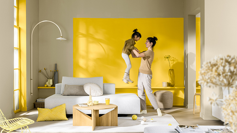 在一个现代起居空间里，妈妈抱起孩子。在她们身后是一面涂刷金盏黄的主题墙。
